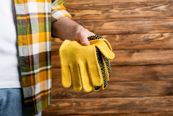 cropped view of handyman in plaid shirt holding work gloves on wooden background, labor day concept - Photo, Image