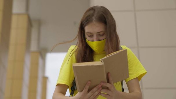 Koulutyttö naamiossa lukee kirjaa seistessään koulussa, suojaa koronavirusta vastaan, covid-19-pandemiaa, ilmansuodatusta maskin suodattimen läpi, modernia koulutusta nuorille. - Valokuva, kuva