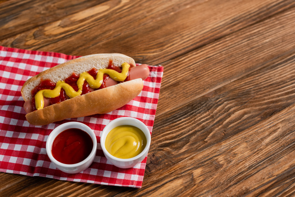 bowls with ketchup and mustard near tasty hot dog on plaid table napkin and wooden surface - Φωτογραφία, εικόνα