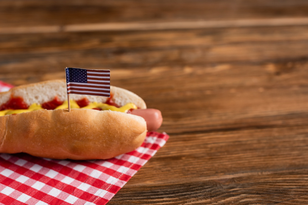 tasty hot dog with small american flag on plaid napkin and wooden table - Фото, изображение