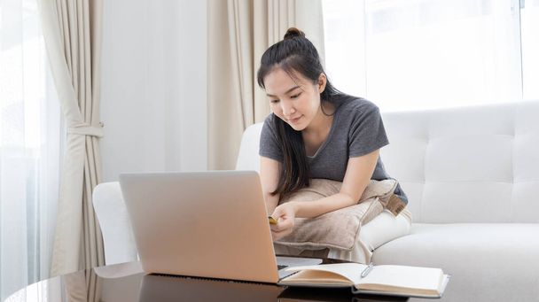 Asiatin sitzt auf Sofa und nutzt Laptop, um online mit Kreditkarte einzukaufen, um sich für Zahlungen oder Online-Transaktionen anzumelden, Finanztransaktionen und Internetsicherheit, Einkaufen mit Kreditkarte. - Foto, Bild