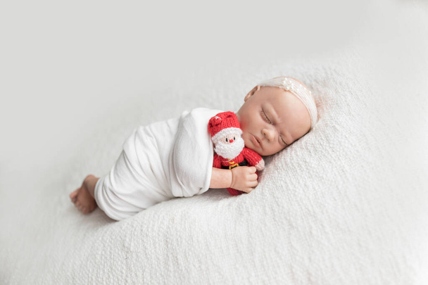 Pasgeboren baby pop gewikkeld in een witte wrap met een witte parelmoer hoofdband en knuffelend een kerstmis kerstman liggend op haar zij op een witte deken. Foto voor baby 's eerste kerst, GEEN MODEL RELEASE VEREIST als model is een herboren pop - Foto, afbeelding
