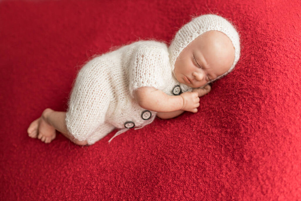 Poupée nouveau-née portant une combinaison en tricot blanc et un bonnet en tricot couché sur le côté sur une couverture rouge. Photo pour bébés premier Noël, AUCUN MODÈLE DE COMMUNIQUÉ REQUIS comme modèle est une poupée renaissante - Photo, image