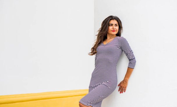 Fashionable portrait of happy beautiful Hispanic Latino girl in striped dress with long hair at city street. Positive emotions. Lifestyle concept, charming dark-haired Arabic woman - Foto, Imagen
