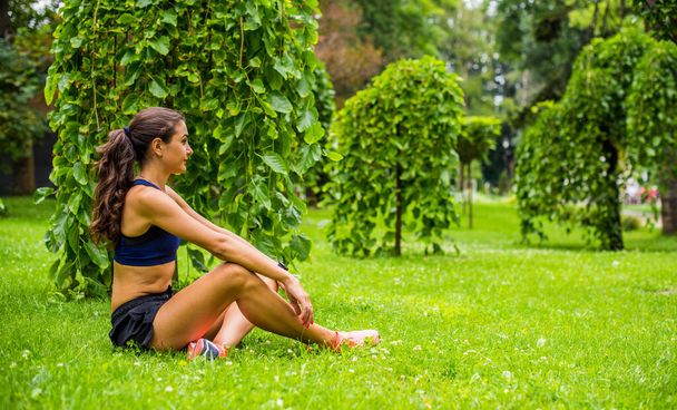 Mooi brunette vrouwelijk fitness meisje buiten aan het trainen in een groen en groen stadspark. Dame in sport top joggen in het park. Gezonde meisje levensstijl achtergrond met kopieerruimte - Foto, afbeelding