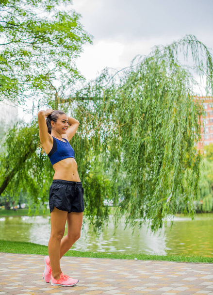 Bela menina de fitness feminina morena se exercitando fora em um parque da cidade frondosa e verde. Senhora em esportes top jogging no parque. Fundo de estilo de vida menina saudável com espaço de cópia - Foto, Imagem