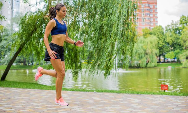 Beautiful brunette female fitness girl exercising outside in a leafy and green city park. Lady in sports top jogging in park. Healthy girl lifestyle background with copy space - Fotó, kép