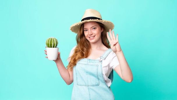 ruda głowa ładna rolnik kobieta uśmiecha się i wygląda przyjaźnie, pokazując numer cztery i trzymając kaktusa - Zdjęcie, obraz