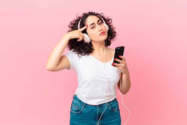pretty arab woman feeling confused and puzzled, showing you are insane with headphones and a smartphone - Фото, изображение