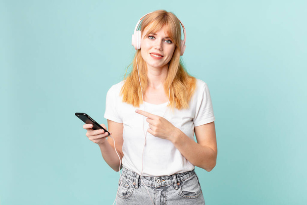 pretty red head woman smiling cheerfully, feeling happy and pointing to the side and listening music with headphones - Foto, Imagen