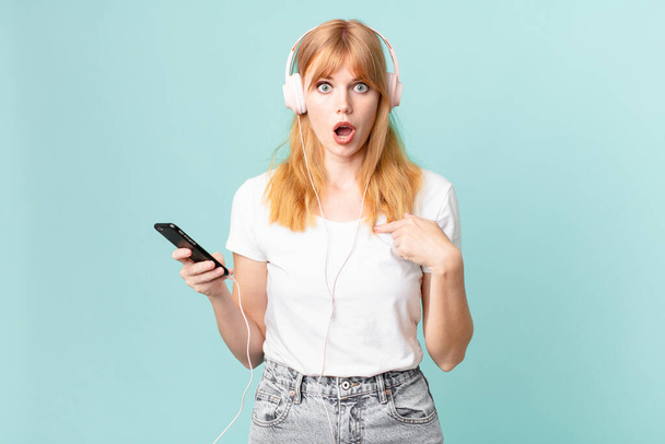 pretty red head woman feeling happy and pointing to self with an excited and listening music with headphones - 写真・画像