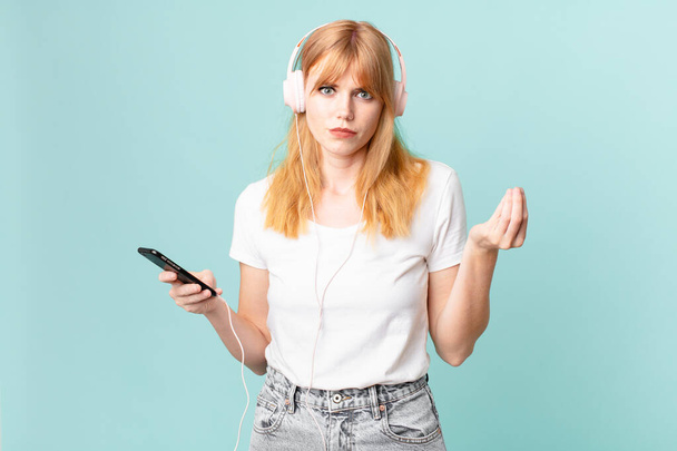 pretty red head woman making capice or money gesture, telling you to pay and listening music with headphones - Foto, afbeelding