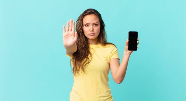 joven bonita mujer buscando seria mostrando abierta palma haciendo stop gesture y sosteniendo un teléfono inteligente - Foto, imagen