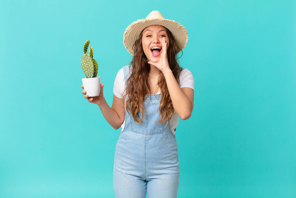 junge hübsche Frau, die sich glücklich fühlt, mit Händen neben dem Mund schreit und einen Kaktus in der Hand hält - Foto, Bild