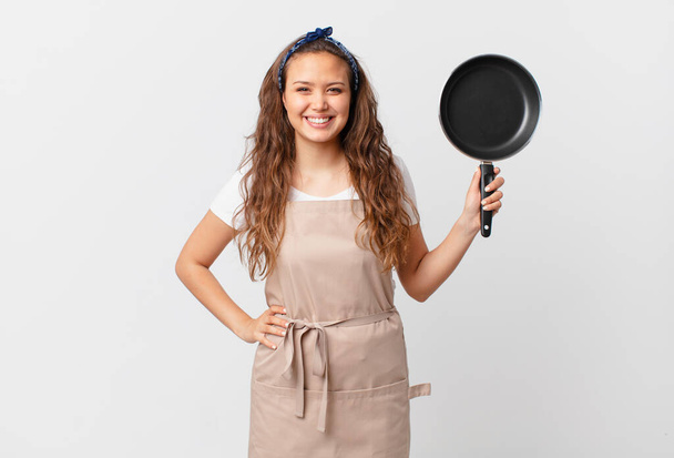 young pretty woman smiling happily with a hand on hip and confident chef concept and holding a pan - Photo, image