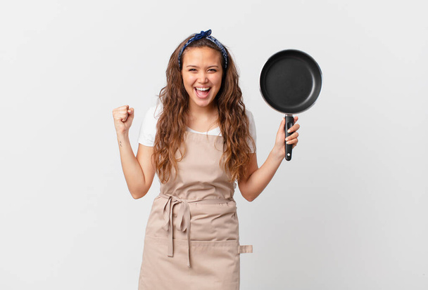 young pretty woman feeling shocked,laughing and celebrating success chef concept and holding a pan - Photo, image