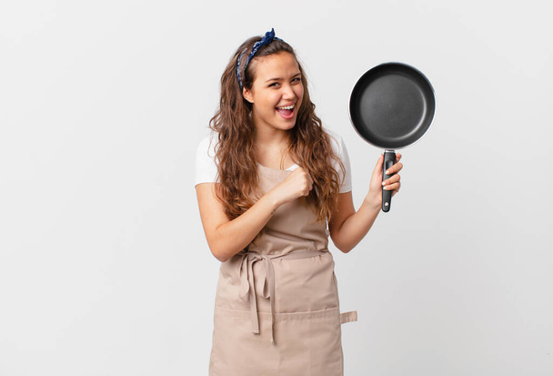 young pretty woman feeling happy and facing a challenge or celebrating chef concept and holding a pan - Foto, immagini