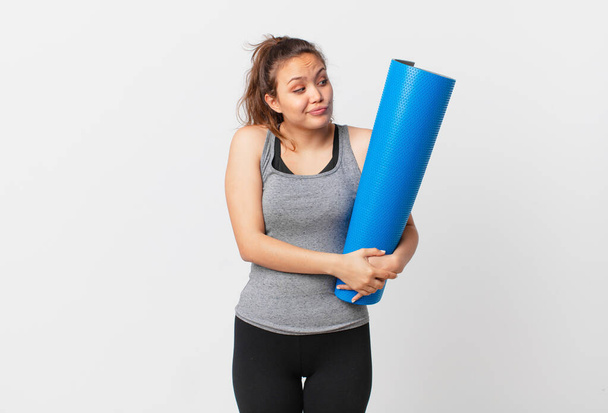 young pretty woman shrugging, feeling confused and uncertain and holding a yoga mat - Fotoğraf, Görsel