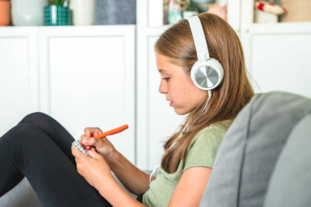 Ένα μικρό κορίτσι με ακουστικά κάθεται στον καναπέ στο σαλόνι και γράφοντας σε ένα σημειωματάριο κατά τη διάρκεια της online εκπαίδευσης στο σπίτι - Φωτογραφία, εικόνα