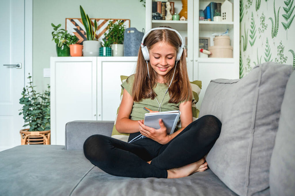 Ein kleines Mädchen mit Kopfhörern sitzt auf dem Sofa im Wohnzimmer und schreibt während des Online-Unterrichts zu Hause in ein Notizbuch - Foto, Bild