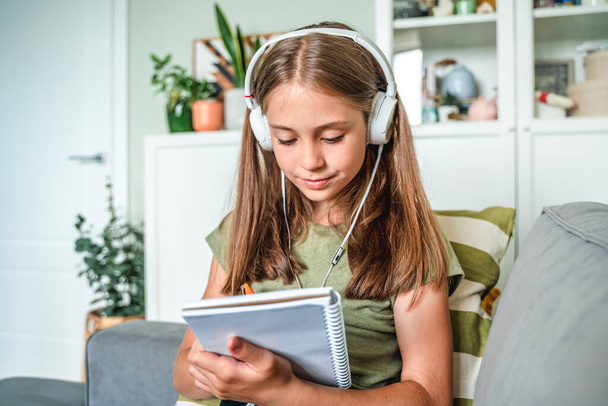 A little girl with headphones is sitting on the sofa in the living room and writing in a notebook during online education at home - Fotografie, Obrázek