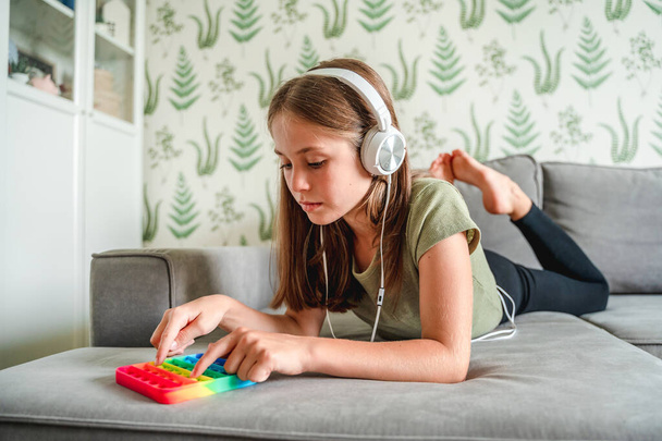 Ein kleines Teenager-Mädchen liegt auf der Couch, hört Musik und entspannt sich mit Hilfe eines Pop-It-Spielzeugs - Foto, Bild