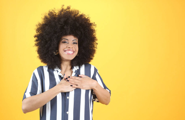 afro kobieta z dłońmi na sercu, szczęśliwy uśmiech, żółte tło - Zdjęcie, obraz