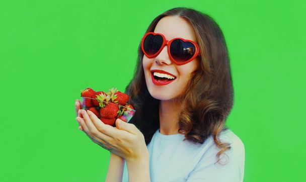 Portrait close up of happy smiling woman with handful of fresh strawberries wearing a red heart shaped sunglasses on green background - Foto, immagini