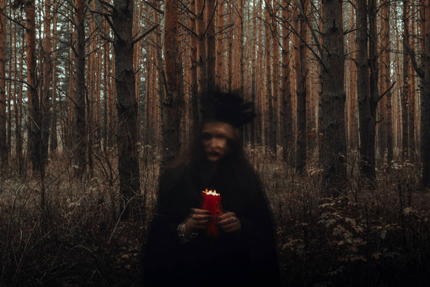 Czarownica w czarnym kostiumie ze świecami w ręku wykonuje rytuał okultystyczny i mistyczne zaklęcia w ciemnym lesie - Zdjęcie, obraz