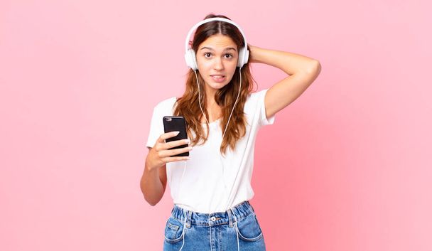 young pretty woman feeling stressed, anxious or scared, with hands on head with headphones and a smartphone - 写真・画像