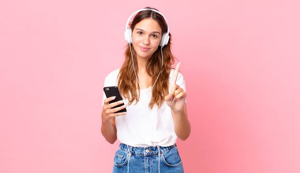 young pretty woman smiling proudly and confidently making number one with headphones and a smartphone - Foto, Imagen