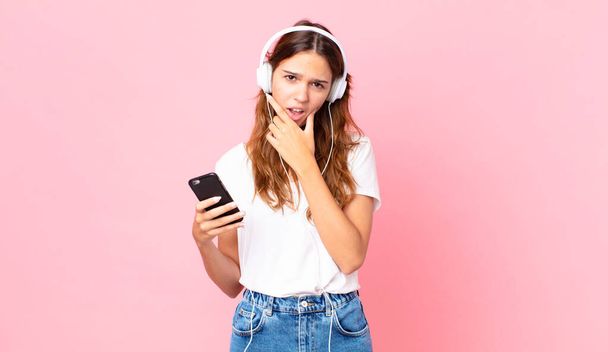 young pretty woman with mouth and eyes wide open and hand on chin with headphones and a smartphone - 写真・画像