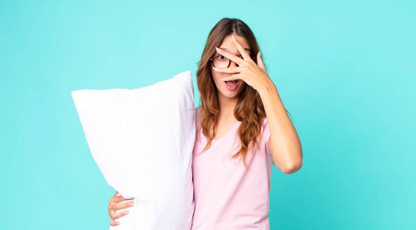young pretty woman looking shocked, scared or terrified, covering face with hand wearing pajamas and holding a pillow - Foto, imagen