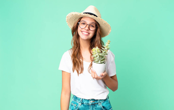 junge hübsche Frau lächelt glücklich mit einer Hand auf der Hüfte und zuversichtlich mit einem Strohhut und hält einen Kaktus - Foto, Bild