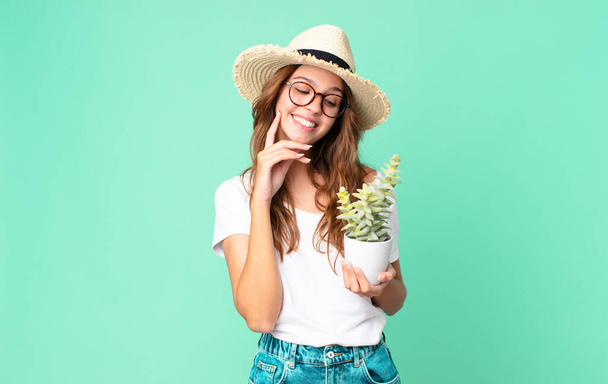 młoda piękna kobieta uśmiechająca się szczęśliwie i marząca na jawie lub wątpiąca w słomkowy kapelusz i trzymająca kaktusa - Zdjęcie, obraz