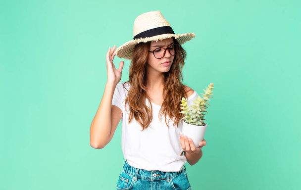 young pretty woman feeling puzzled and confused, scratching head with a straw hat and holding a cactus - Fotoğraf, Görsel