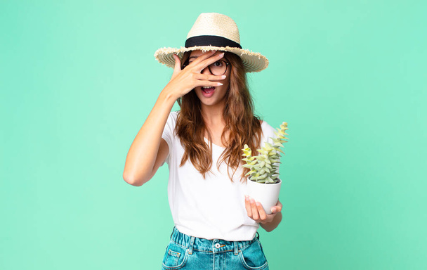 young pretty woman looking shocked, scared or terrified, covering face with hand with a straw hat and holding a cactus - Φωτογραφία, εικόνα
