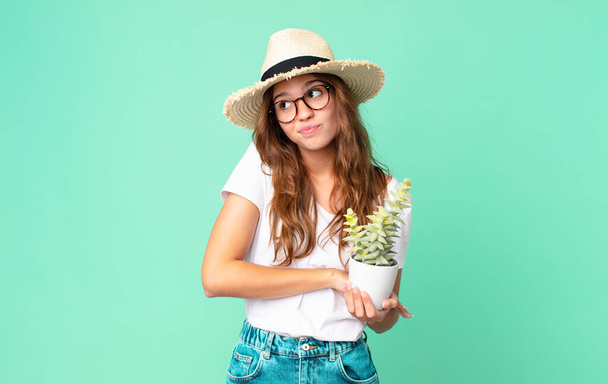 young pretty woman shrugging, feeling confused and uncertain with a straw hat and holding a cactus - Фото, зображення