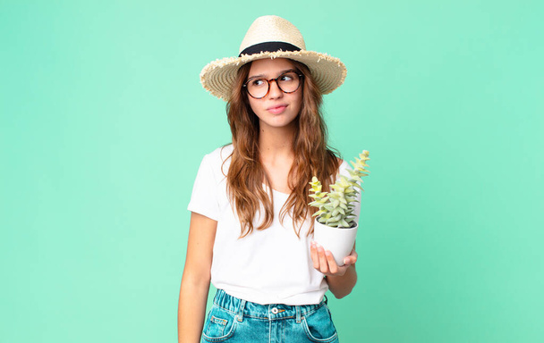 young pretty woman feeling sad, upset or angry and looking to the side with a straw hat and holding a cactus - Photo, Image
