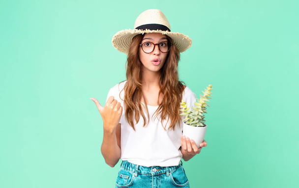 young pretty woman looking astonished in disbelief with a straw hat and holding a cactus - Photo, Image