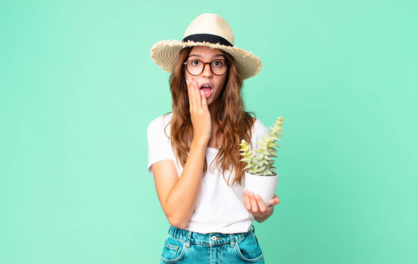 young pretty woman feeling shocked and scared with a straw hat and holding a cactus - Fotografie, Obrázek