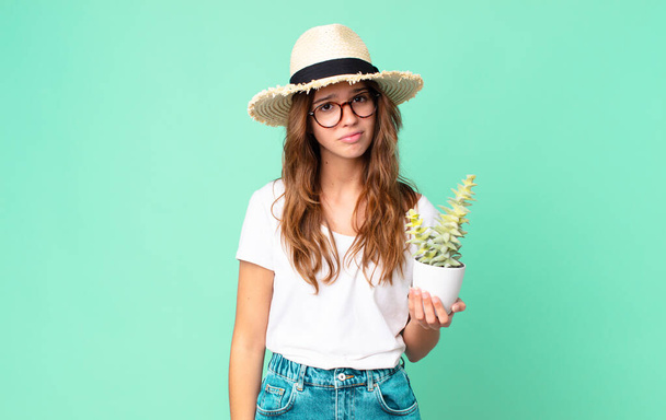 young pretty woman feeling sad and whiney with an unhappy look and crying with a straw hat and holding a cactus - Foto, Imagen