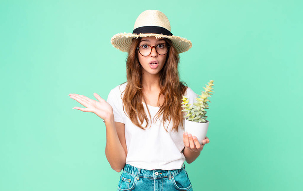 young pretty woman looking surprised and shocked, with jaw dropped holding an object with a straw hat and holding a cactus - Foto, Bild