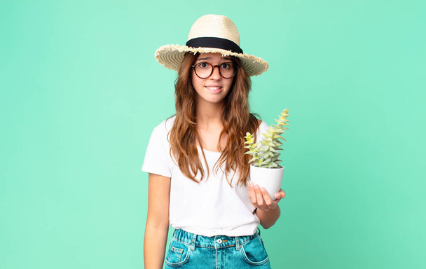 young pretty woman looking puzzled and confused with a straw hat and holding a cactus - Foto, afbeelding