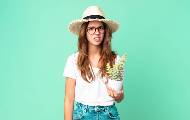 young pretty woman feeling puzzled and confused with a straw hat and holding a cactus - Foto, afbeelding