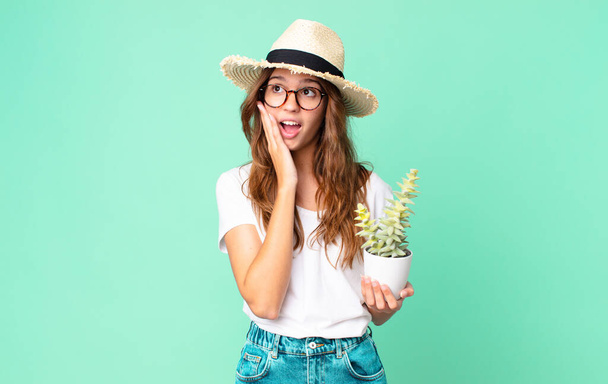 young pretty woman feeling happy, excited and surprised with a straw hat and holding a cactus - Foto, immagini