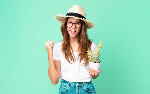 young pretty woman shouting aggressively with an angry expression with a straw hat and holding a cactus - Фото, зображення