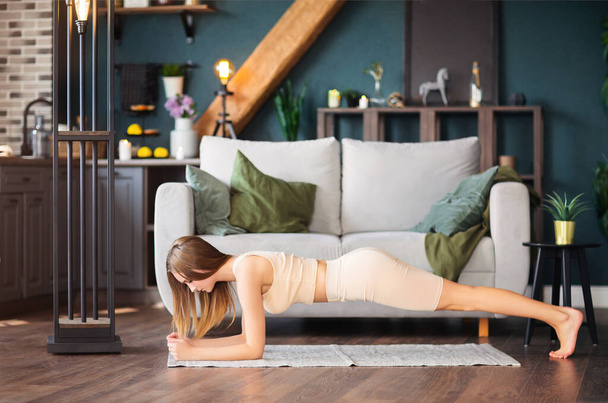 Стройная молодая женщина в спортивной одежде отходит в сторону, практикуя йогу возле дивана дома - Фото, изображение