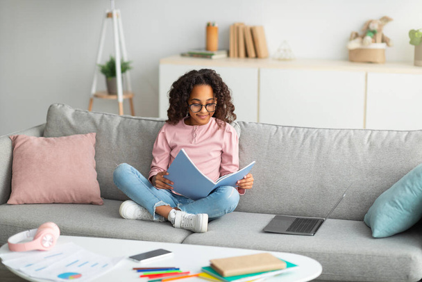 Концепція навчання та виховання. Усміхнена афроамериканська дівчина читає і робить домашнє завдання, сидячи на дивані з ноутбуком
 - Фото, зображення