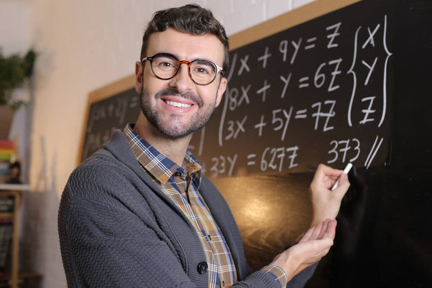 primer plano retrato de guapo joven profesor escribir ejemplo de matemáticas en pizarra en el aula - Foto, Imagen